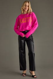 Woman wearing a bright pink sweater with 'Merry' text and black pants on a gray background