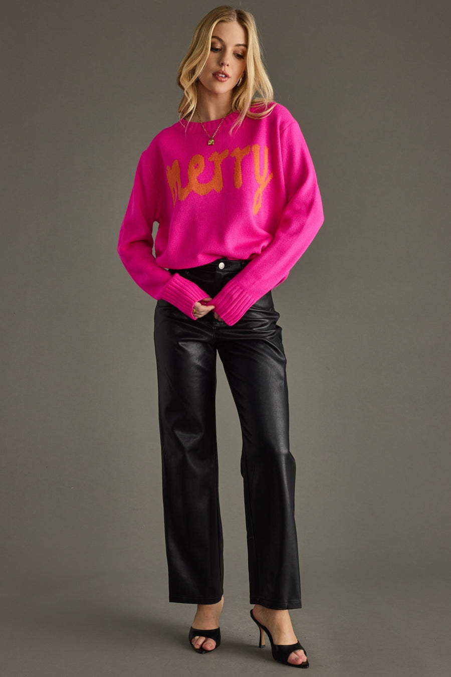 Woman wearing a bright pink sweater with 'Merry' text and black pants on a gray background