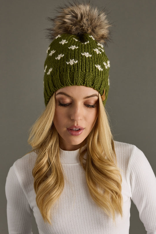 Woman wearing a green knit beanie with a fur pom-pom against a gray background