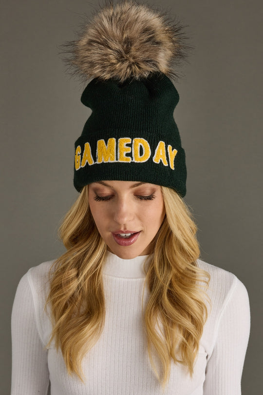 Green beanie with 'GAME DAY' text and fur pom-pom worn by a person against a gray background