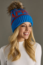 Woman wearing a blue knit beanie with a fur pom-pom against a gray background