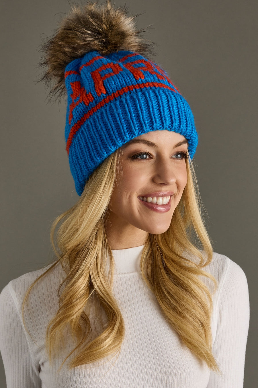 Woman wearing a blue knit beanie with a fur pom-pom against a gray background