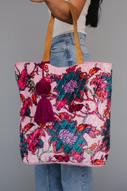 A person holds the Amara Floral Tote from Panache Apparel Collection, featuring leather suede straps, colorful floral embroidery, and two purple tassels. They are dressed in blue jeans and a white top.