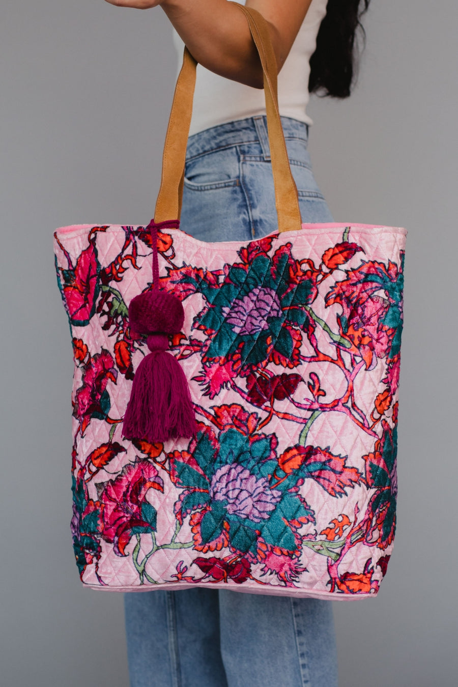 A person holds the Amara Floral Tote from Panache Apparel Collection, featuring leather suede straps, colorful floral embroidery, and two purple tassels. They are dressed in blue jeans and a white top.