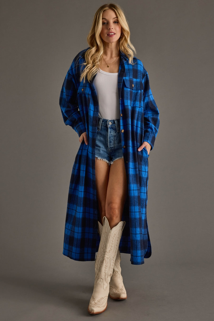 Woman wearing a blue plaid coat over a white top and denim shorts, standing against a gray background.
