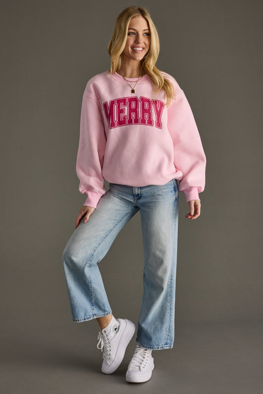 Woman wearing a pink sweatshirt with 'Merry' text, blue jeans, and white sneakers on a gray background