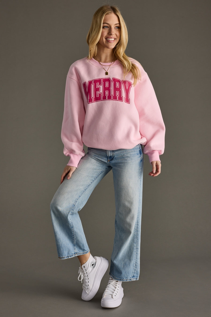 Woman wearing a pink sweatshirt with 'Merry' text, blue jeans, and white sneakers on a gray background