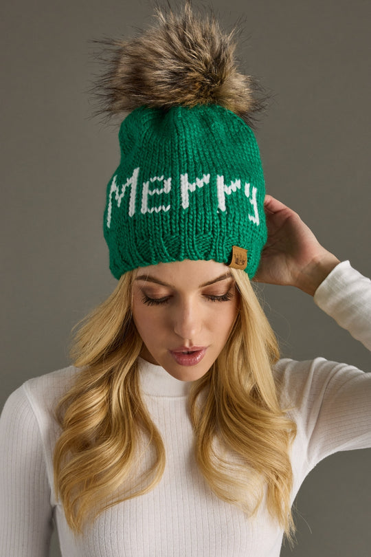 Green knit beanie with 'Merry' text and fur pom-pom worn by a person against a neutral background
