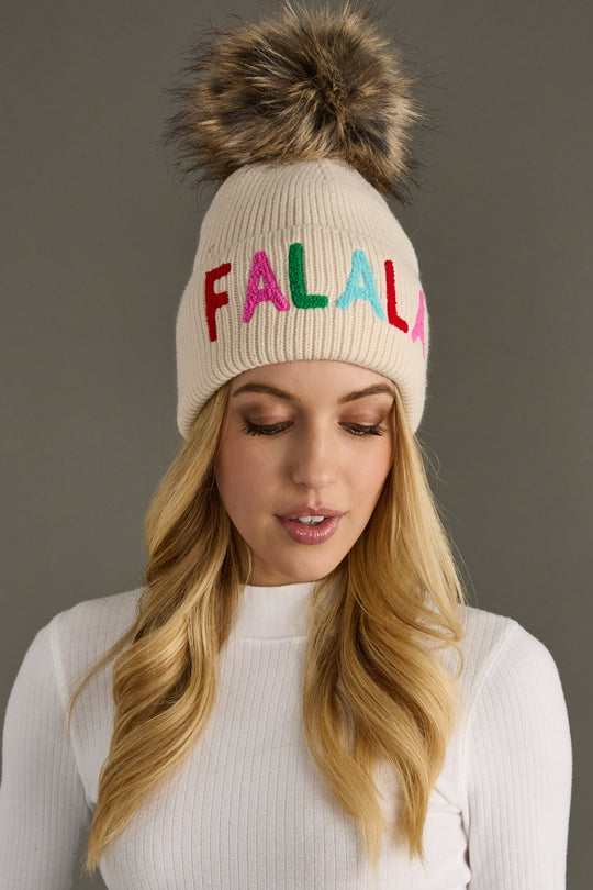 Beige knit beanie with colorful 'FALALA' letters and fur pom-pom worn by a model against a gray background