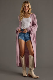 Woman wearing a long pink cardigan, white top, denim shorts, and beige cowboy boots on a gray background