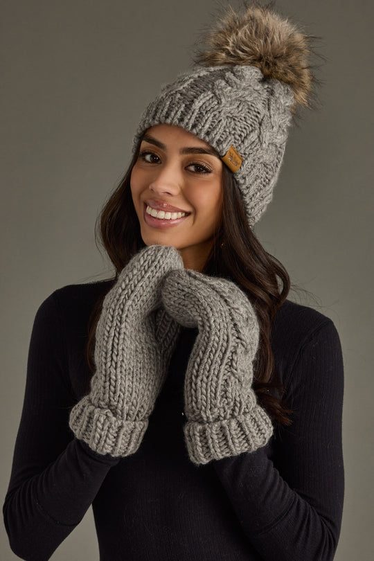 Woman wearing a gray knit beanie with a pom-pom and matching mittens against a neutral background