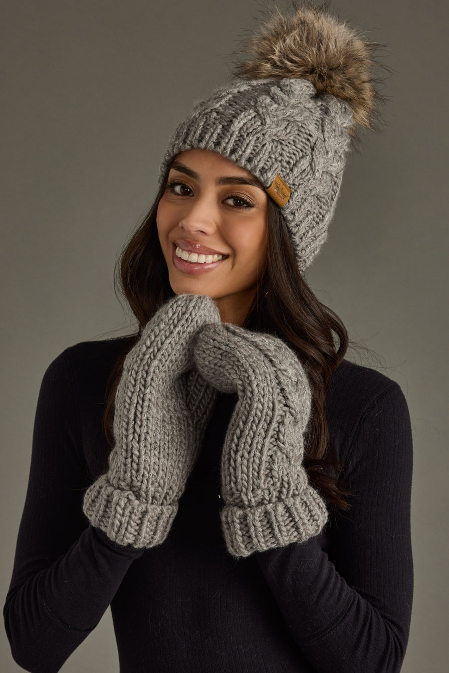 Woman wearing a gray knit beanie with a pom-pom and matching mittens against a neutral background