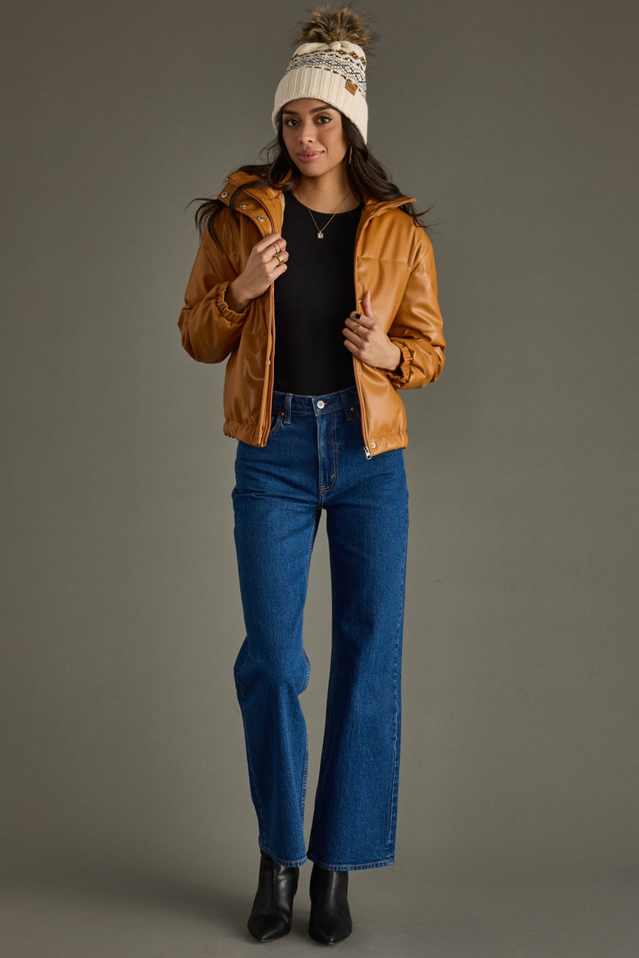 Woman wearing a brown jacket, black top, blue jeans, and a white beanie against a gray background