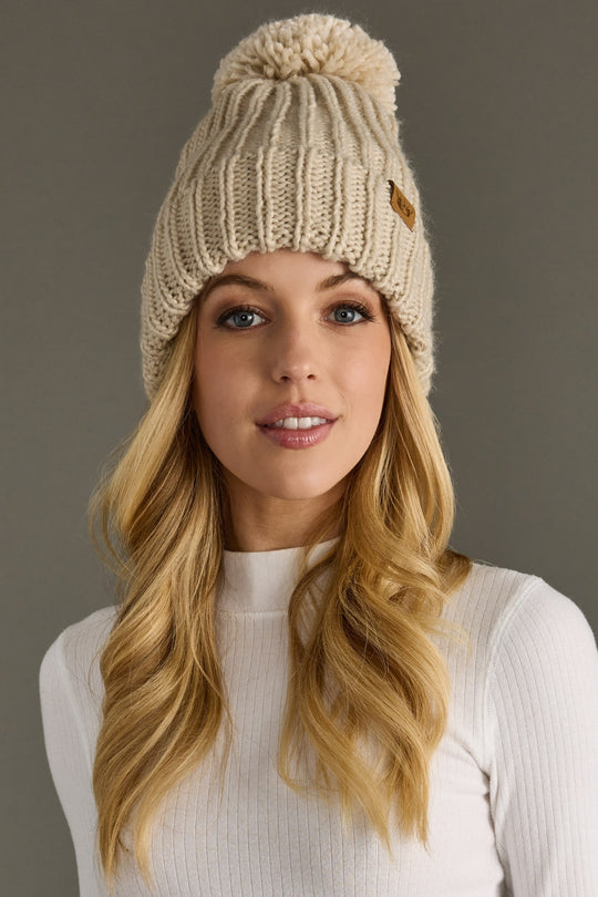 Woman wearing a beige knitted beanie with a pom-pom against a gray background