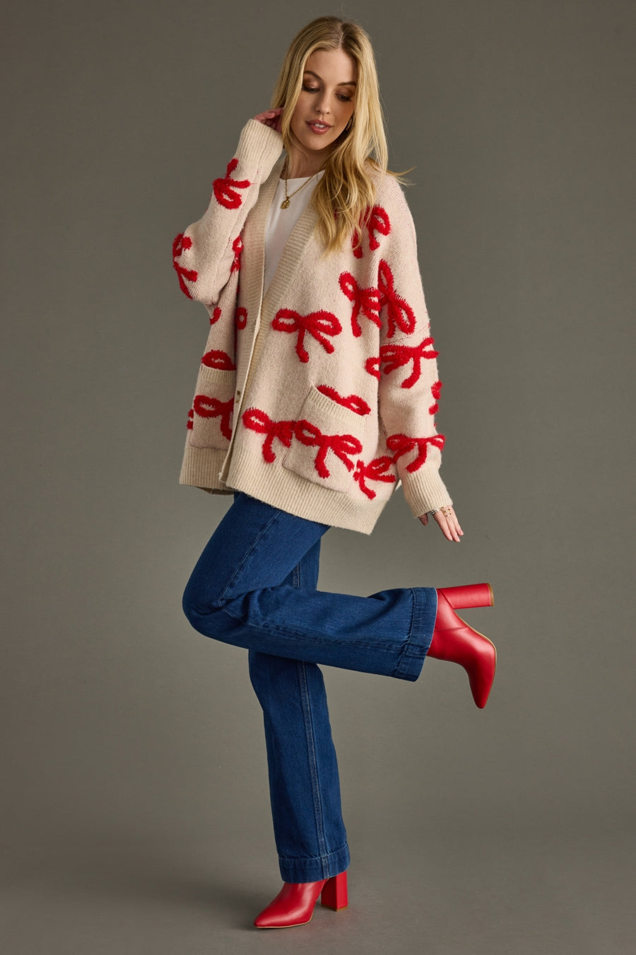 Woman wearing a beige sweater with red patterns, blue jeans, and red shoes on a gray background