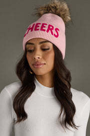 Woman wearing a pink 'CHEERS' beanie with a fur pom-pom on a gray background