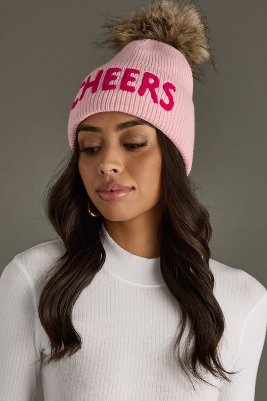 Woman wearing a pink 'CHEERS' beanie with a fur pom-pom on a gray background