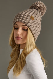 Woman wearing a taupe knitted beanie with a pom-pom on a gray background