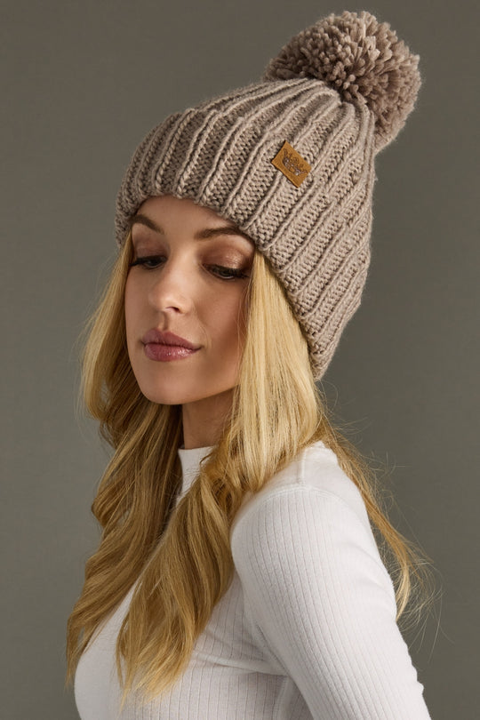 Woman wearing a taupe knitted beanie with a pom-pom on a gray background