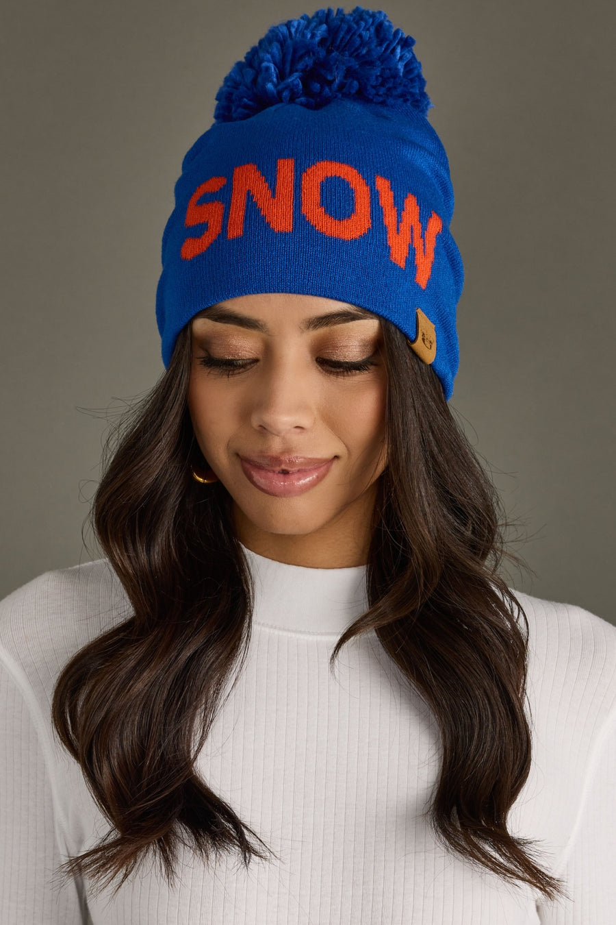 A woman with long dark hair wears the Panache Apparel Collection Snow Pom Hat in royal blue, featuring bold red "SNOW" text. She pairs it with a white ribbed sweater and looks down with a slight smile.