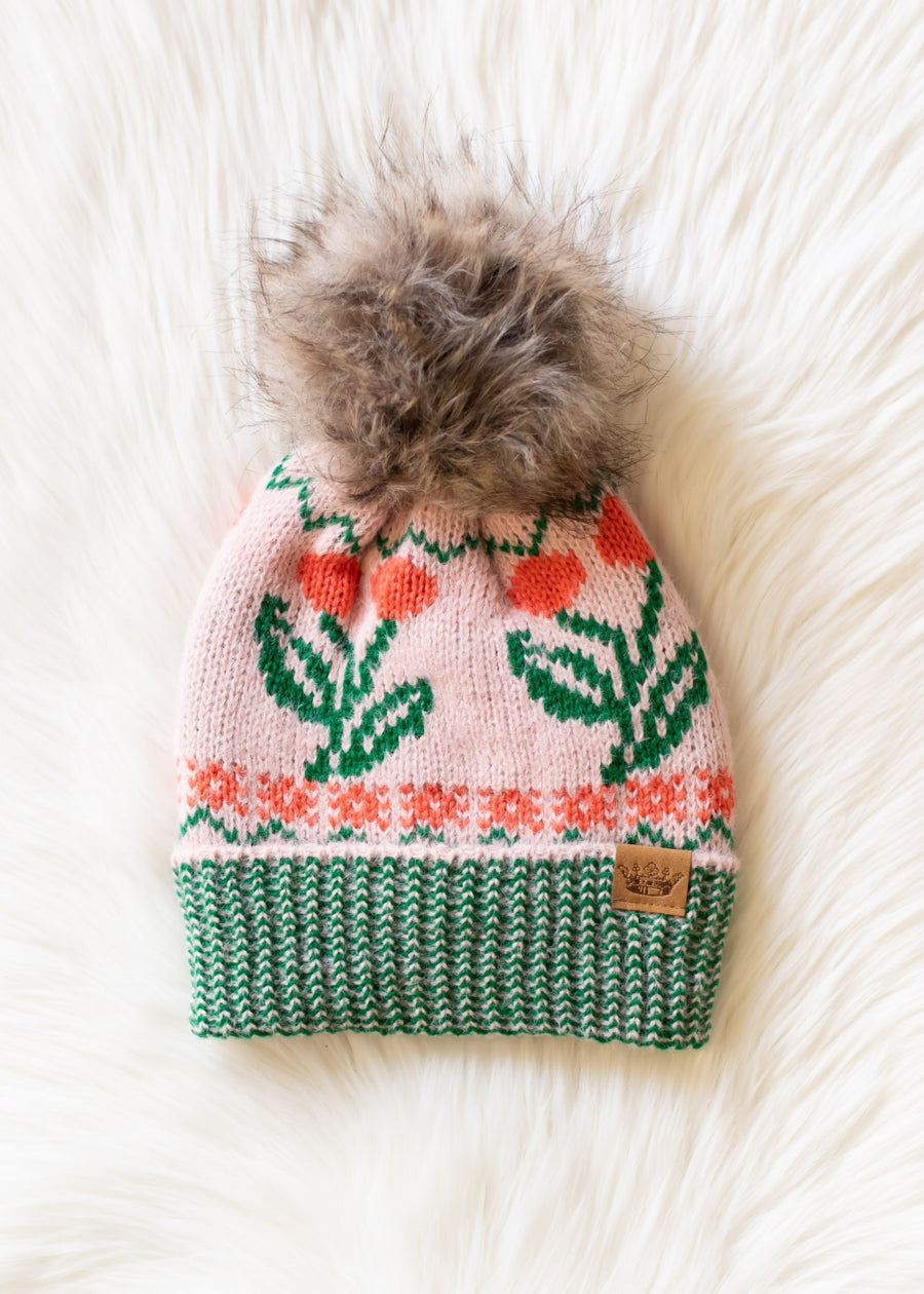The Cherry Pattern Pom Hat - Pink by Panache Apparel Collection features a pink floral knit design with a green and white ribbed brim, orange accents, fleece lining for warmth, and a large faux fur pom, shown on a white furry surface.