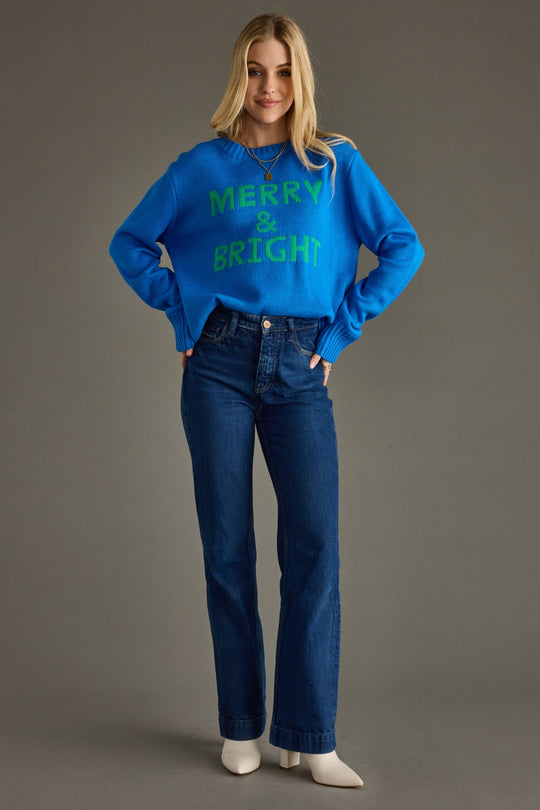 Woman wearing a blue sweater with 'Merry & Bright' text and blue jeans on a gray background