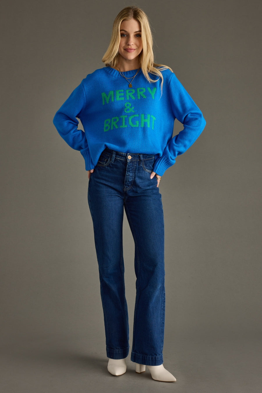 Woman wearing a blue sweater with 'Merry & Bright' text and blue jeans on a gray background