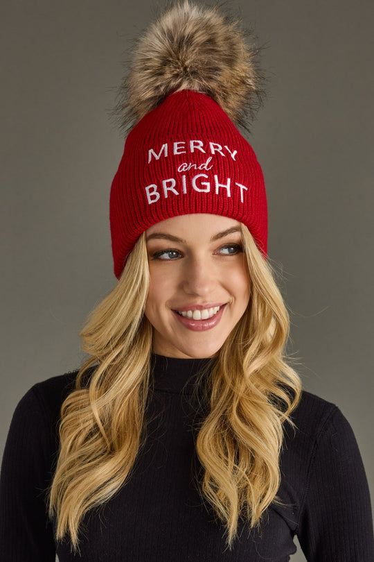 Woman wearing a red beanie with 'Merry and Bright' text and a fur pom-pom on a gray background