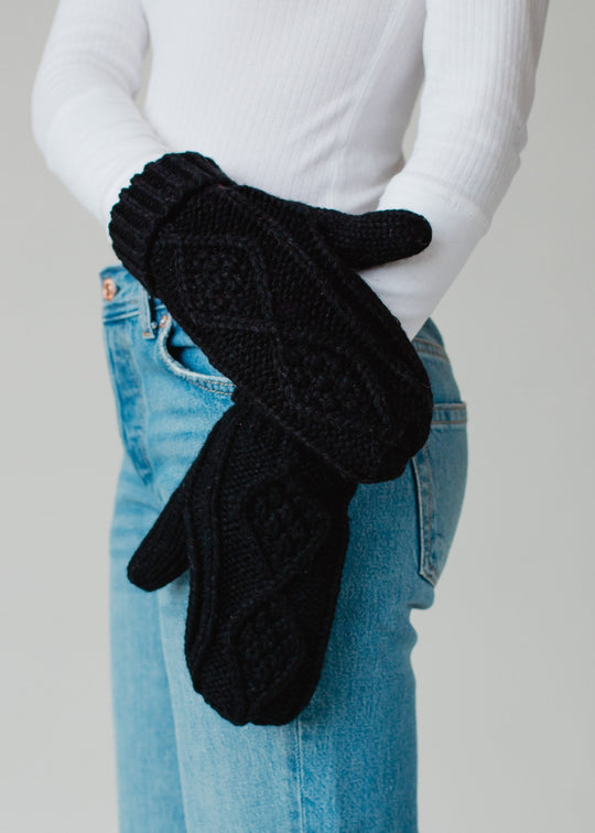 Black knitted gloves worn by a person with a white shirt and blue jeans on a gray background