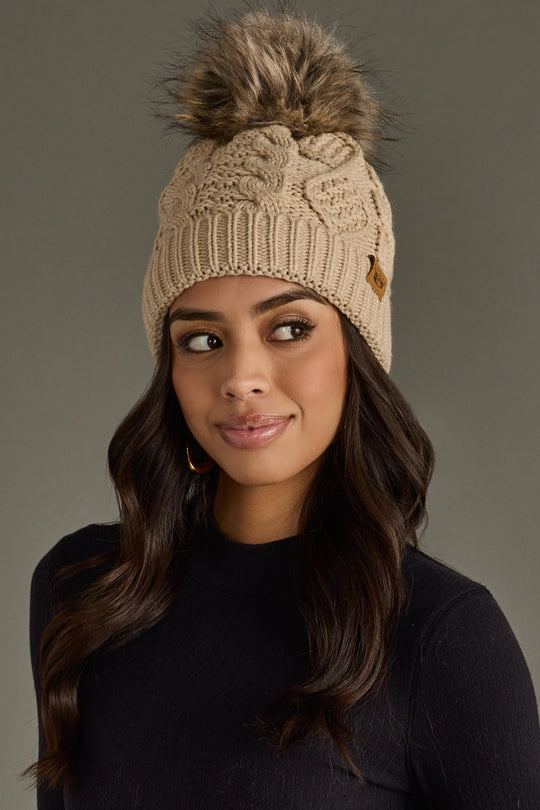Woman wearing a beige knit beanie with a pom-pom against a gray background