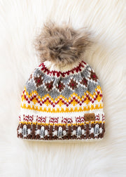 The Xenea Pom Hat by Panache Apparel Collection is a colorful, fleece-lined knit hat with a faux fur pom-pom and a white, yellow, gray, red, and brown pattern, shown on a soft white furry background.