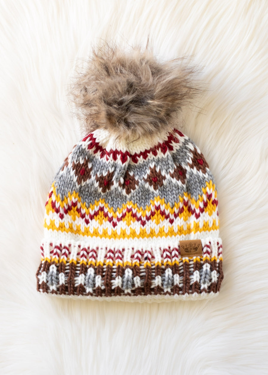 The Xenea Pom Hat by Panache Apparel Collection is a colorful, fleece-lined knit hat with a faux fur pom-pom and a white, yellow, gray, red, and brown pattern, shown on a soft white furry background.