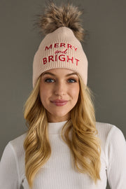 Woman wearing a beige knit hat with 'Merry and Bright' text and a fur pom-pom on a gray background