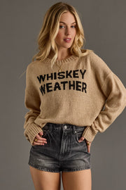 A cozy tan knit sweater featuring bold black “Whiskey Weather” lettering across the front for a fun, fall-ready statement. Designed with a relaxed, easy fit and a soft textured knit, it includes a classic crew neckline, long sleeves, and ribbed cuffs and hem for comfortable everyday wear. Perfect for layering or wearing on its own, this sweater pairs effortlessly with denim for a warm, casual seasonal look.