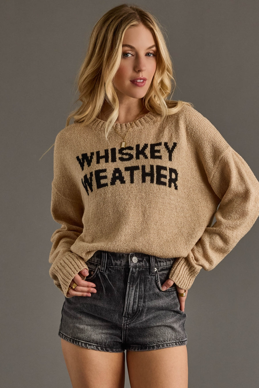 A cozy tan knit sweater featuring bold black “Whiskey Weather” lettering across the front for a fun, fall-ready statement. Designed with a relaxed, easy fit and a soft textured knit, it includes a classic crew neckline, long sleeves, and ribbed cuffs and hem for comfortable everyday wear. Perfect for layering or wearing on its own, this sweater pairs effortlessly with denim for a warm, casual seasonal look.