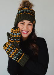 A smiling woman models the Panache Apparel Collection Reyna Pom Hat in dark green with fleece lining and faux fur pom, paired with matching patterned mittens in green, yellow, and white against a light gray background.