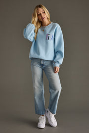 Woman wearing a light blue sweatshirt with a colorful design, light blue jeans, and white sneakers on a gray background