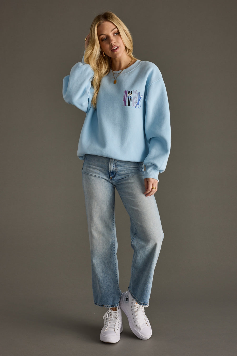 Woman wearing a light blue sweatshirt with a colorful design, light blue jeans, and white sneakers on a gray background