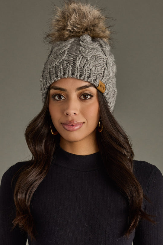 Woman wearing a gray knit beanie with a fur pom-pom against a neutral background
