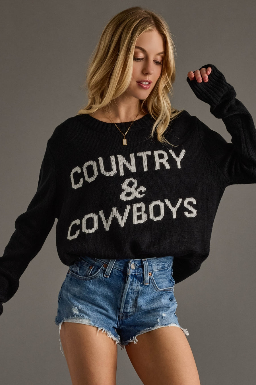 Woman wearing a black sweater with 'COUNTRY & COWBOYS' text and denim shorts on a gray background