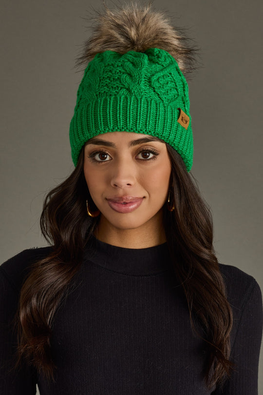 Woman wearing a green knitted beanie with a fur pom-pom on a gray background