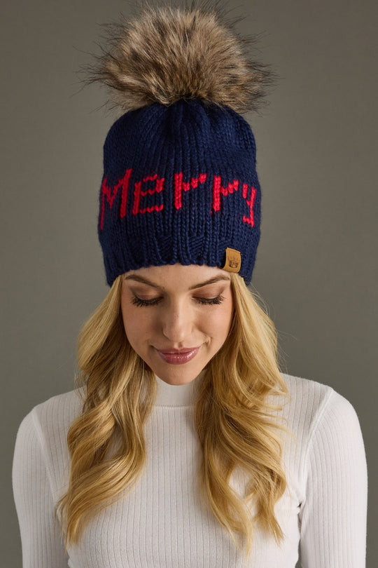 Navy blue knit beanie with red 'Merry' text and fur pom-pom worn by a person against a gray background