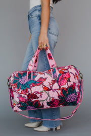 A woman in jeans and a white top holds the Amara Floral Duffel by Panache Apparel Collection, featuring pink handles, a removable strap, and a zipper closure, standing against a plain gray background.