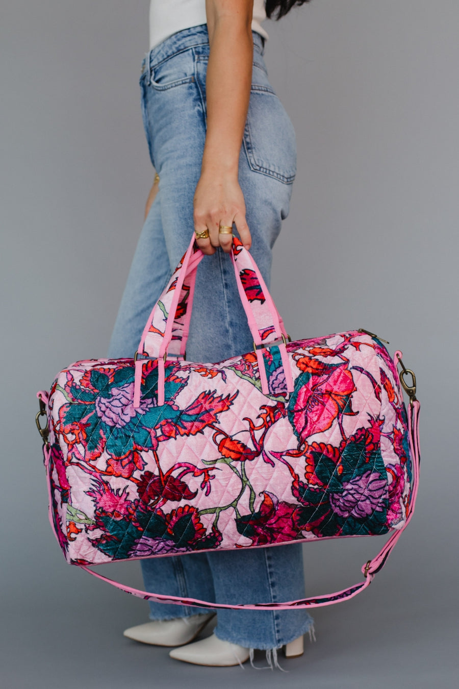 A woman in jeans and a white top holds the Amara Floral Duffel by Panache Apparel Collection, featuring pink handles, a removable strap, and a zipper closure, standing against a plain gray background.