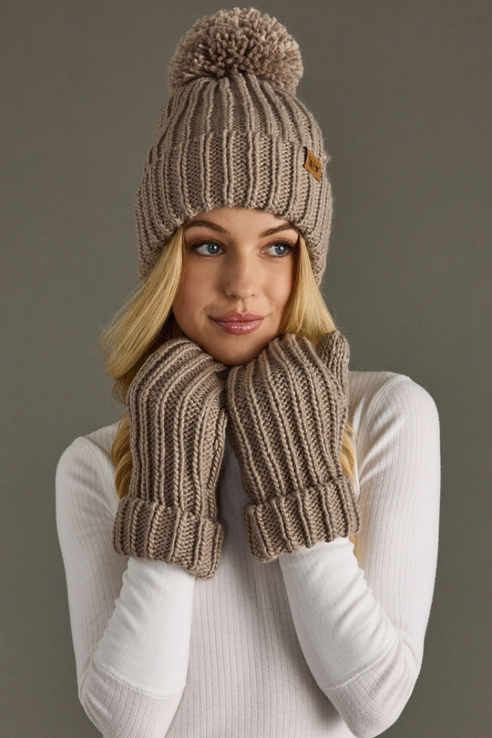 Woman wearing a taupe knit hat with pom-pom and mittens on a gray background