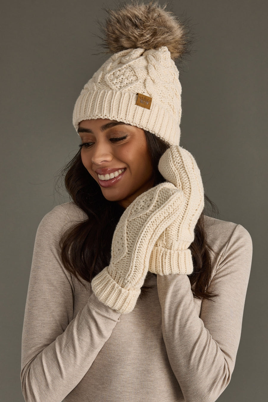 Woman wearing a cream knit beanie and mittens against a gray background