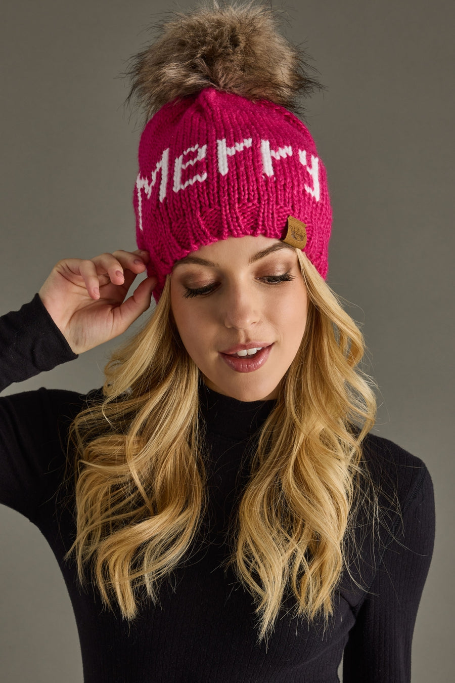 Person wearing a pink knit hat with a fur pom-pom and 'Merry' text on a gray background