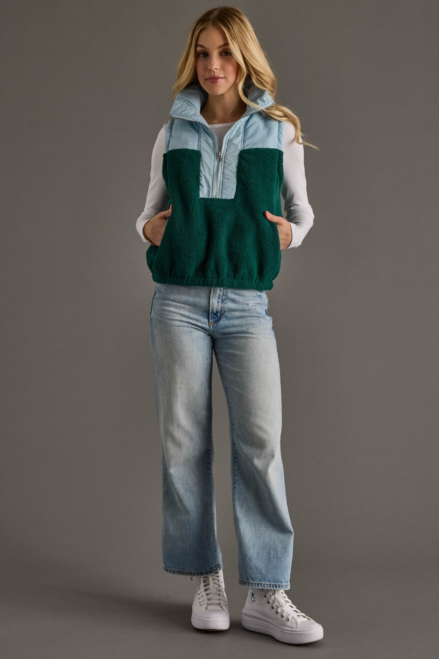 A woman stands against a gray background wearing the Panache Apparel Collection Light Blue & Green Vest over a light blue long-sleeve shirt, paired with light blue jeans and white sneakers, with her hands in her vest pockets.
