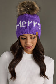 A woman wears the Panache Apparel Collection Merry Pom Hat - Purple, a fleece-lined knit with "Merry" in white, paired with a white ribbed turtleneck top, standing against a plain gray background.