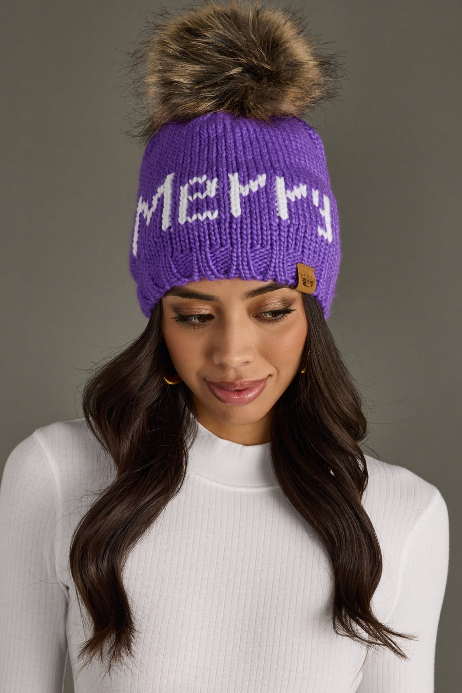 A woman wears the Panache Apparel Collection Merry Pom Hat - Purple, a fleece-lined knit with "Merry" in white, paired with a white ribbed turtleneck top, standing against a plain gray background.